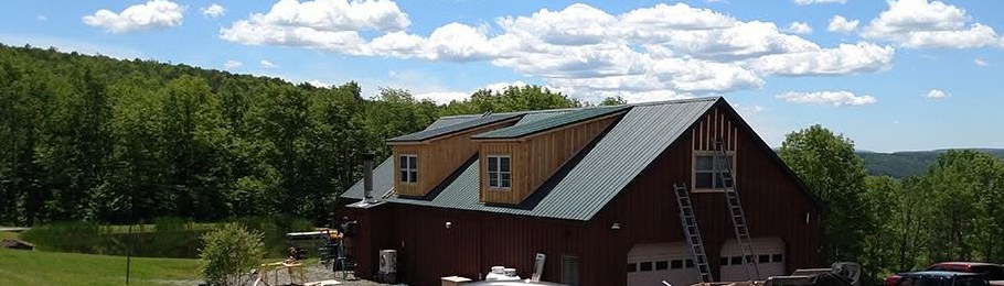 A hilltop garage and barn clad in dark board-and-batten with a green standing-seam metal roof, two shed dormers, and ladders up the gable end